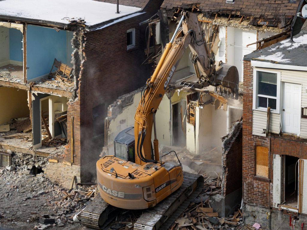An excavator demolishes an old brick building at a construction site, creating debris and dust.