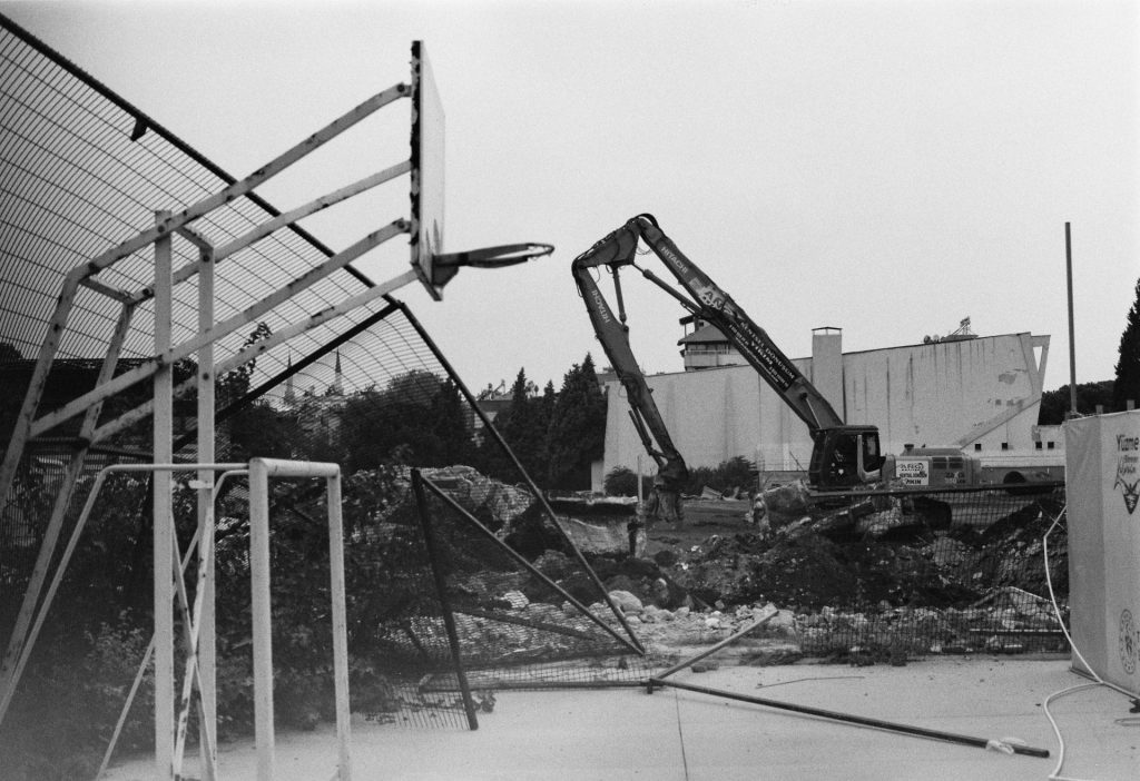 Black and white image of an urban demolition site featuring a dismantled basketball hoop and heavy machinery.